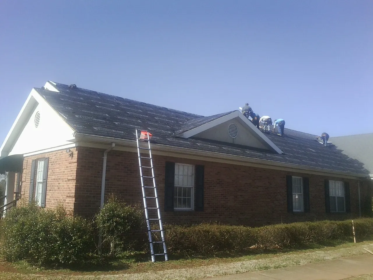 Crew tearing shingles off a brick ranch roof with a ladder set — Metal Roof Repair work by our crew in Roland