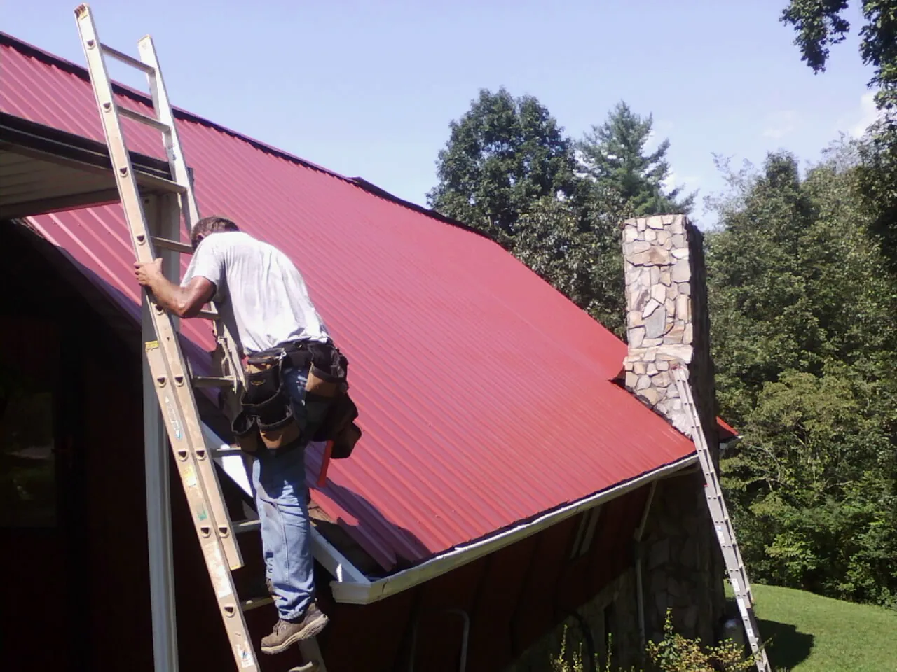 Roofer on a ladder beside a red metal roof and stone chimney — Soffit & Fascia Repair by Roofing Brothers in Roland